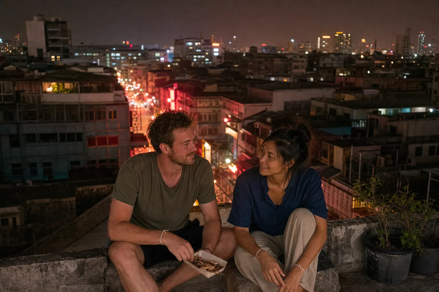 Alex and Jey on a rooftop above Bangkok’s Chinatown at night, the city’s neon and traffic below, a paper tray of grilled squid between them